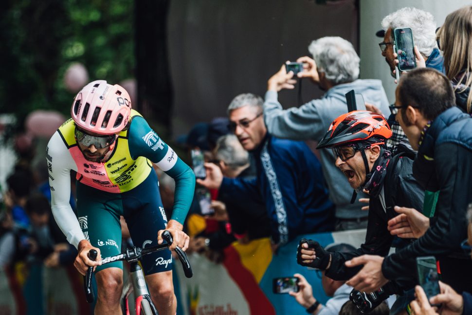 Cyclist getting roared on by crowd in pink helmet and yellow, pink and green kit